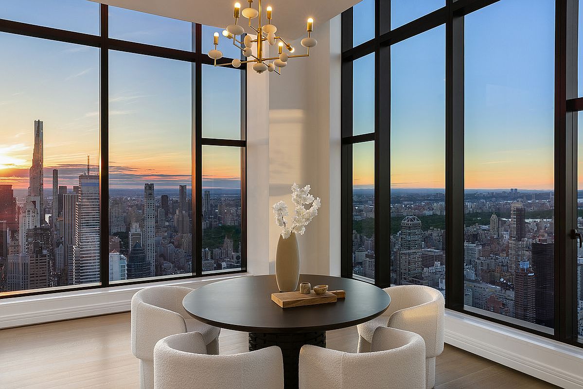 This sophisticated dining area features a sleek, dark circular table surrounded by four plush, cream-colored upholstered chairs, centered beneath a modern, multi-armed chandelier. The room is defined by floor-to-ceiling windows that offer a breathtaking, panoramic view of the New York City skyline at sunset. The minimalist decor, including a simple vase with white blossoms, emphasizes the luxury and height of this high-rise residence.