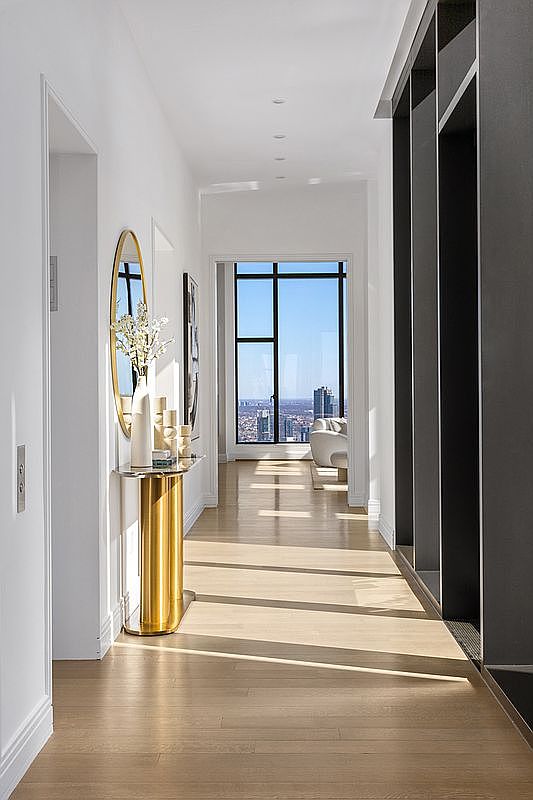 This elegant, sun-drenched hallway features light hardwood flooring and a striking gold pedestal console table topped with a decorative vase and mirror. The space is framed by a dramatic, dark-toned architectural wall feature on the right, leading the eye toward a floor-to-ceiling window that offers a breathtaking, high-rise urban cityscape view. The minimalist design and clean lines create a sophisticated, airy atmosphere typical of a luxury modern apartment.