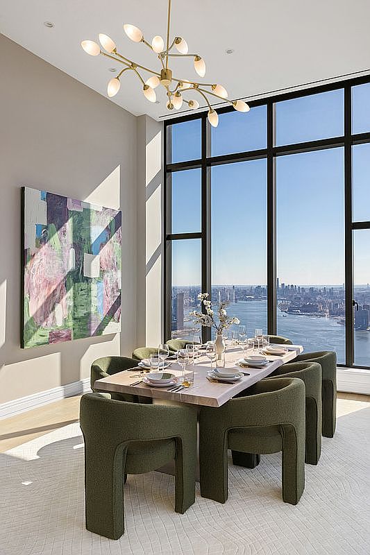 This sophisticated dining room features a minimalist wooden table surrounded by six plush, olive-green upholstered chairs, set against a backdrop of floor-to-ceiling windows offering a breathtaking panoramic view of the city skyline and river. A striking, modern brass chandelier with delicate, leaf-like glass shades hangs above the table, while a large, vibrant abstract painting adds a pop of color to the neutral-toned wall. The space exudes a sense of refined luxury and calm, perfectly capturing the essence of high-end urban living.