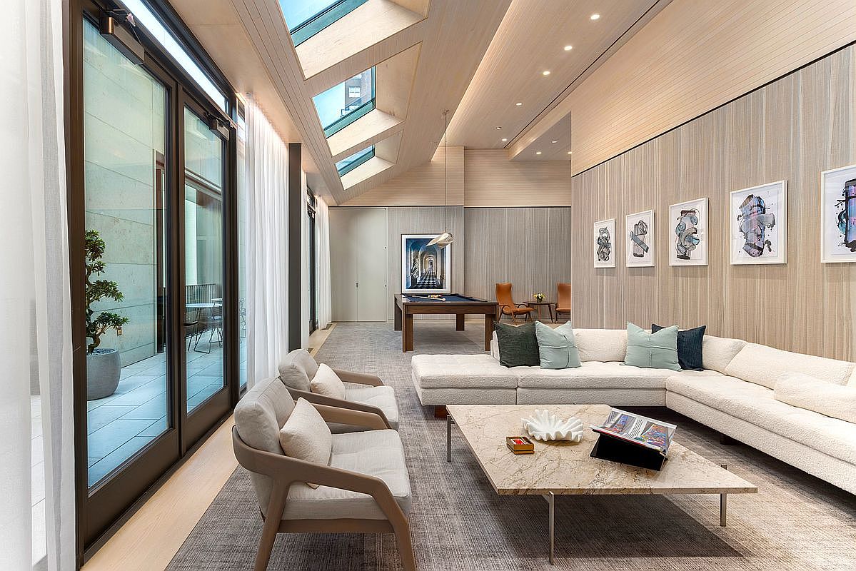 This sophisticated, modern living space features a neutral color palette, high-end finishes, and a seamless transition to an outdoor terrace through floor-to-ceiling glass doors. The room is anchored by a large, plush sectional sofa and a marble coffee table, complemented by a pool table in the background and skylights that flood the area with natural light. The overall aesthetic is one of refined luxury, perfect for high-end entertaining.