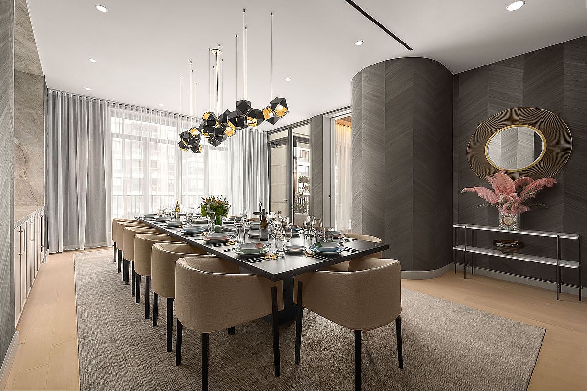 This sophisticated dining room features a long, dark rectangular table surrounded by elegant, light-beige upholstered chairs, set atop a large neutral area rug. A striking, modern geometric chandelier hangs above the table, while a curved wall with dark, textured chevron-patterned wallpaper serves as a dramatic focal point behind a sleek console table and circular mirror. The space is bathed in natural light from floor-to-ceiling windows draped in soft, floor-length curtains, creating an atmosphere of refined luxury and contemporary style.