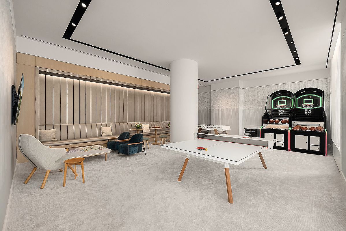 This modern, spacious game room features a sleek white ping-pong table as the centerpiece, complemented by dual basketball arcade games and a foosball table in the background. A comfortable lounge area with plush seating and a long, upholstered bench provides a relaxing space for residents to socialize. The room is brightly lit with recessed lighting and has a clean, contemporary aesthetic perfect for a luxury apartment community.