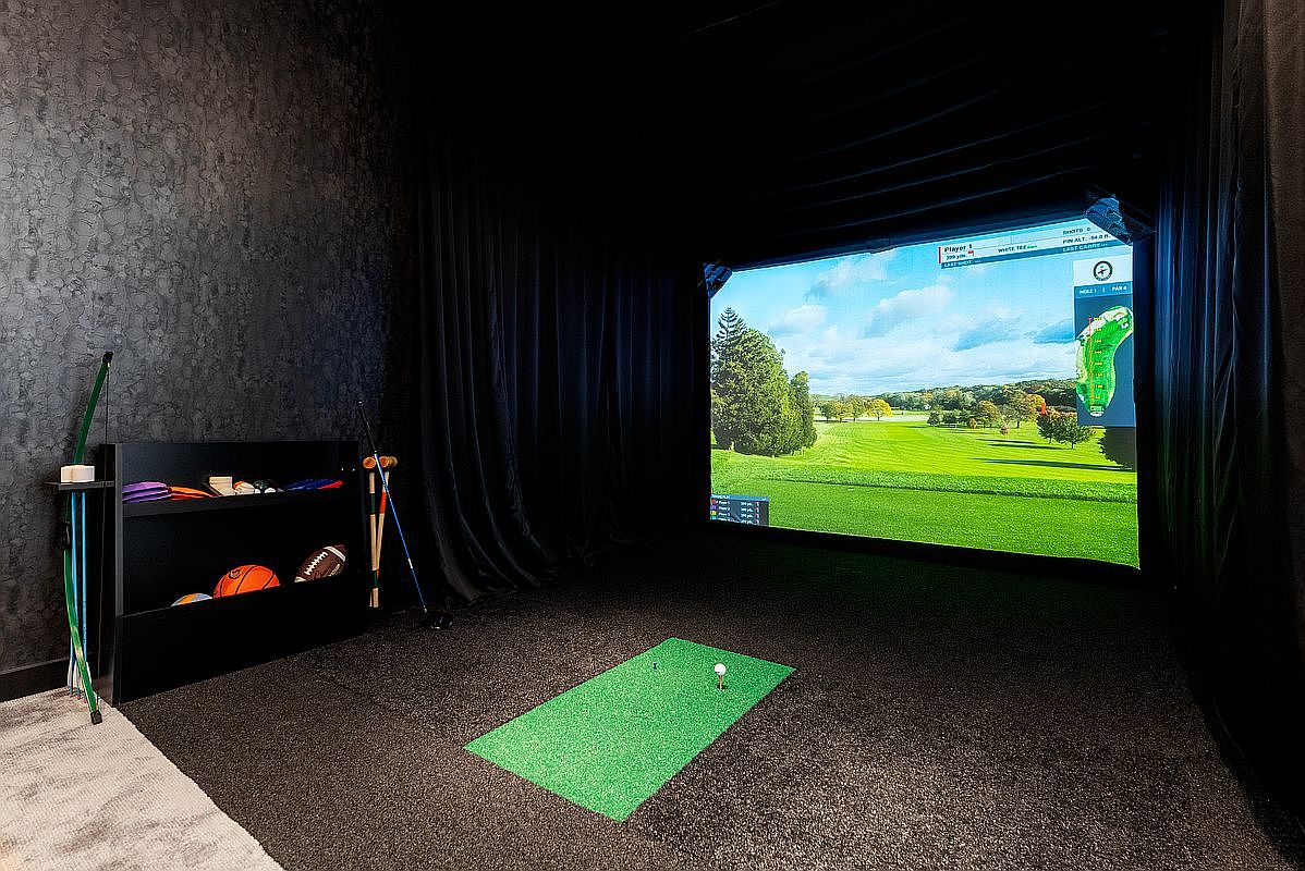 This image showcases a dedicated indoor golf simulator room, featuring a large projection screen displaying a virtual golf course. The space is equipped with a green turf hitting mat, a black storage shelf holding various sports equipment, and dark, sound-dampening curtains surrounding the screen. The room offers a high-end, immersive entertainment experience perfect for sports enthusiasts.