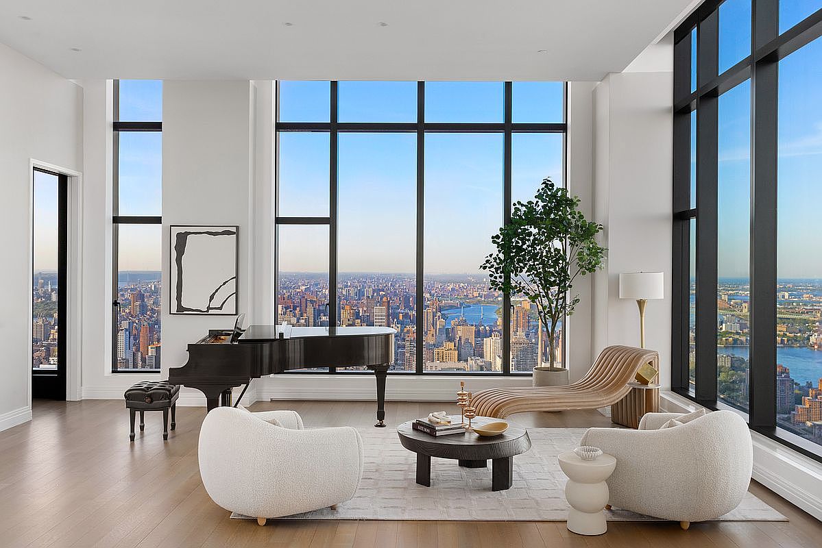 This sophisticated, high-rise living room features floor-to-ceiling windows that offer a breathtaking, panoramic view of the city skyline and river. The space is elegantly furnished with a black grand piano, two plush cream-colored armchairs, and a sculptural wooden chaise lounge arranged around a circular coffee table. The minimalist, modern aesthetic is complemented by light hardwood floors and a neutral color palette, creating an atmosphere of luxury and tranquility.