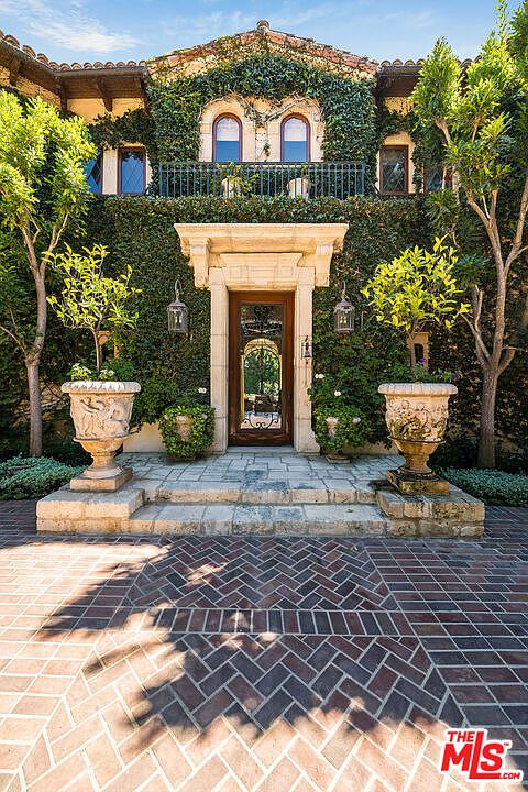 This elegant Mediterranean-style entryway features a grand wooden door framed by ornate stone molding and flanked by two large, decorative stone urns holding small trees. The facade is heavily covered in lush green ivy, creating a classic, timeless aesthetic, while the ground is paved with a sophisticated herringbone-patterned brick. The perspective is a centered, eye-level shot that emphasizes the symmetry and architectural detail of the home's entrance.