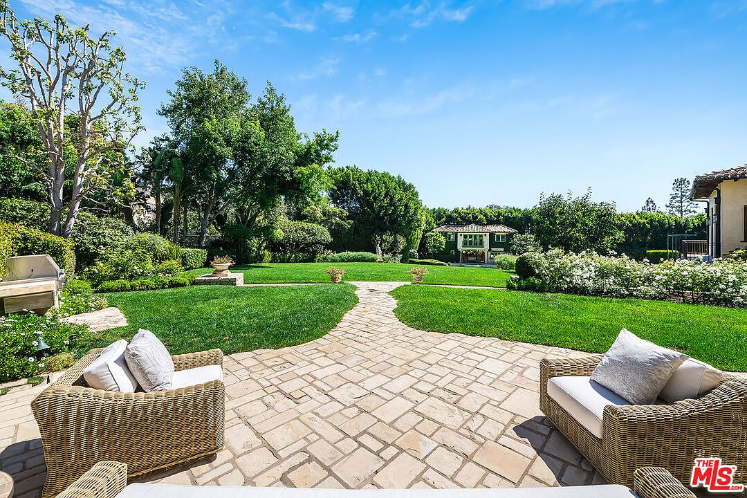 This expansive backyard features a stone-paved patio area furnished with comfortable wicker seating, overlooking a lush, manicured lawn. The garden is framed by mature trees, vibrant green hedges, and blooming white rose bushes, creating a serene and private atmosphere. In the distance, a charming secondary structure with a balcony adds architectural interest to this well-maintained outdoor retreat.