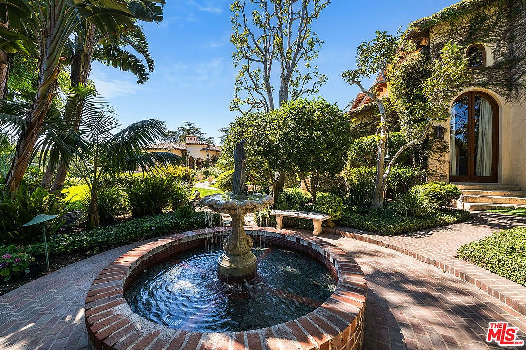This serene garden scene features a classic tiered stone fountain as the central focal point, surrounded by a circular brick patio. Lush greenery, including mature palm trees and manicured shrubs, frames the space, while a stone bench offers a quiet place to sit near the house's exterior wall. The perspective is at eye level, capturing the tranquil atmosphere of a Mediterranean-inspired estate grounds.