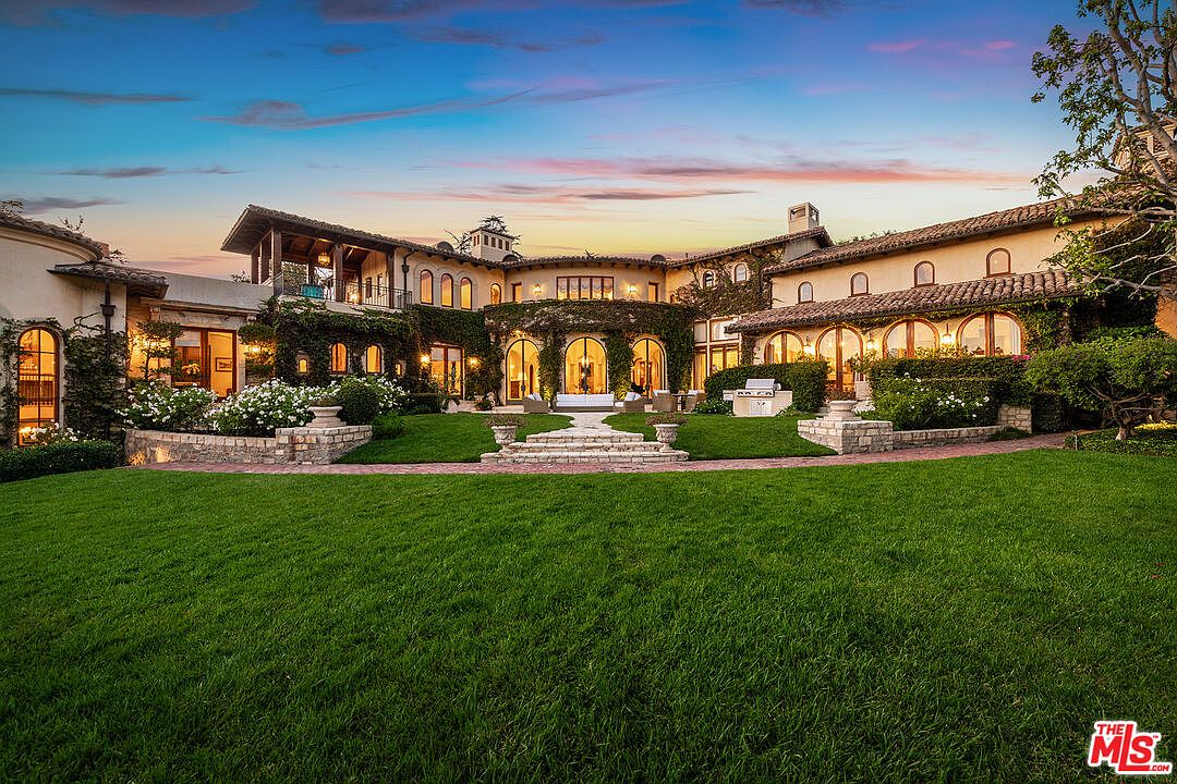 This expansive Mediterranean-style estate is captured from the rear, showcasing a sprawling manicured lawn leading up to a multi-level residence. The home features warm-toned stucco walls, a classic clay tile roof, and numerous arched glass doors that open onto a stone-paved patio area. Lush landscaping, including climbing vines and mature trees, frames the structure, creating a serene and luxurious atmosphere at dusk.