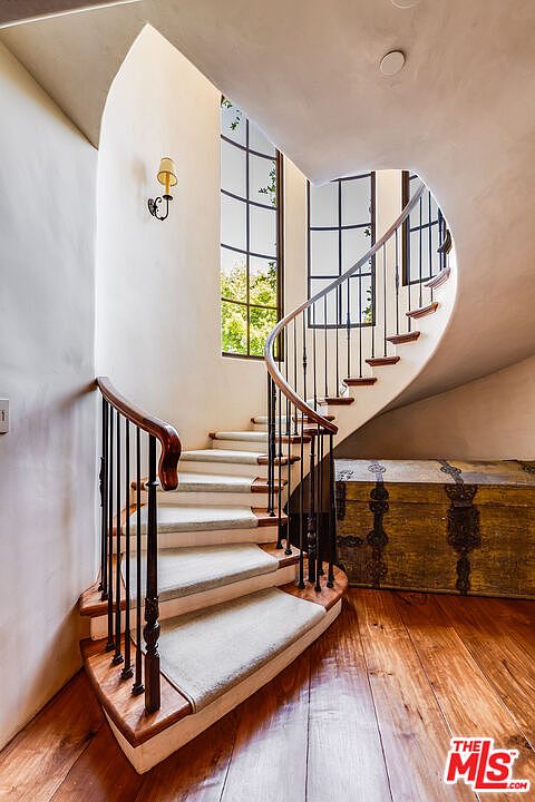 This elegant interior space features a dramatic curved staircase with a dark wood handrail and black metal balusters. The stairs are finished with light-toned treads and a central runner, leading up toward tall, multi-paned windows that allow natural light to fill the area. A large, antique-style wooden chest with ornate metal hardware sits at the base of the stairs on the warm-toned wood flooring, enhancing the classic, sophisticated aesthetic of the entryway.