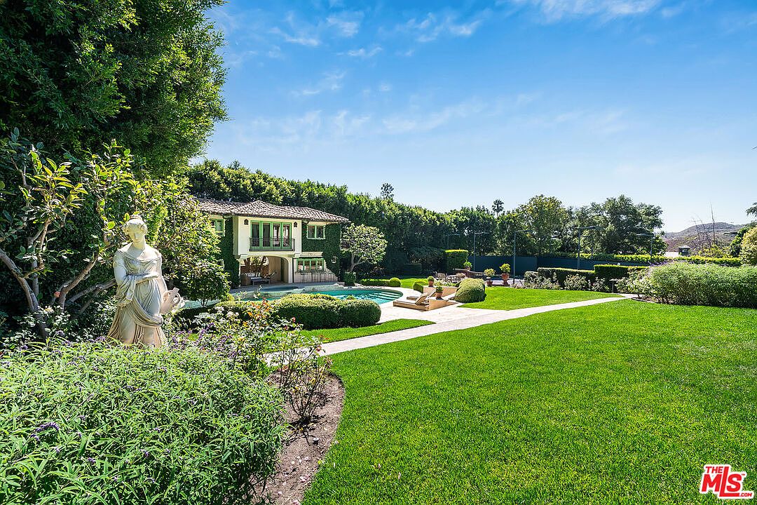 This expansive backyard features a lush, manicured lawn that leads toward a Mediterranean-style home with ivy-covered walls and a swimming pool. A classical-style statue stands prominently in the foreground, surrounded by vibrant greenery and mature trees that provide a sense of privacy. The scene is captured from a ground-level perspective, highlighting the seamless transition between the garden, the pool area, and the residence.