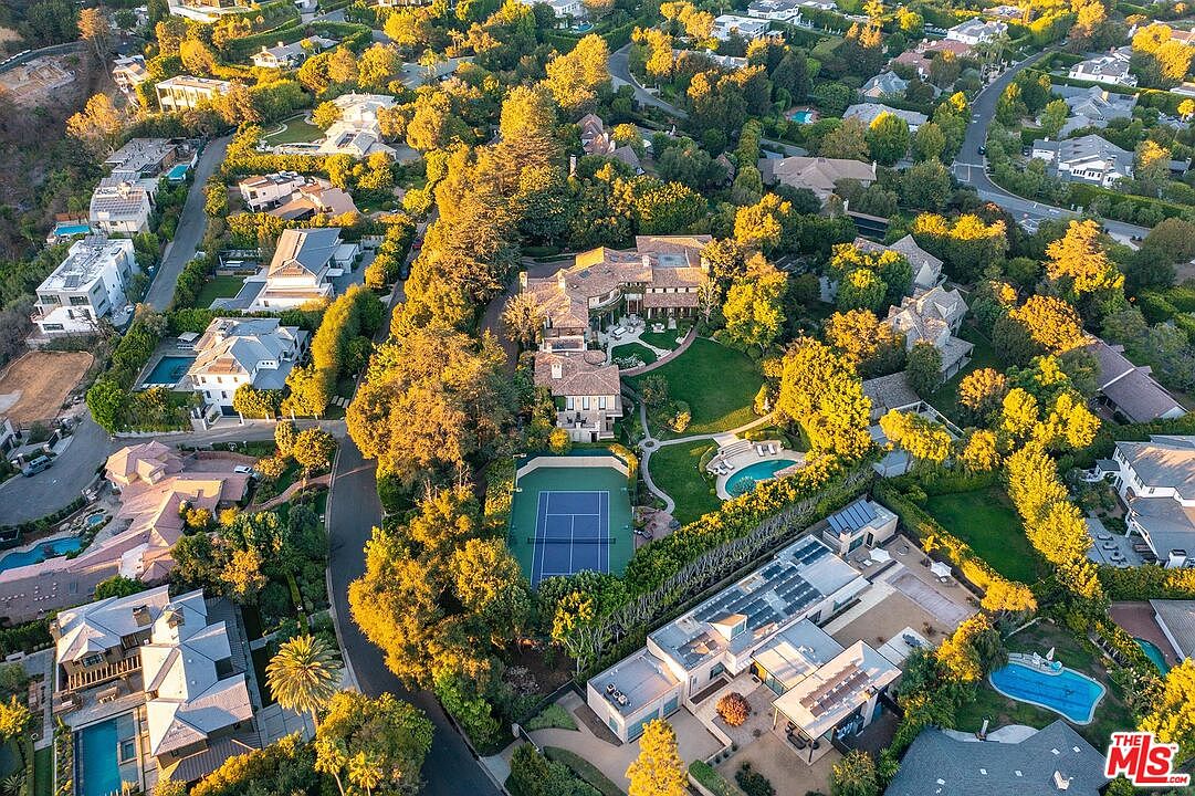 This high-angle aerial view captures a sprawling luxury estate nestled within a lush, tree-lined neighborhood. The property features a large main residence with a complex roofline, a private tennis court, a swimming pool, and expansive manicured lawns. The surrounding area is densely landscaped, showcasing a high-end residential community with winding streets and multiple neighboring homes.