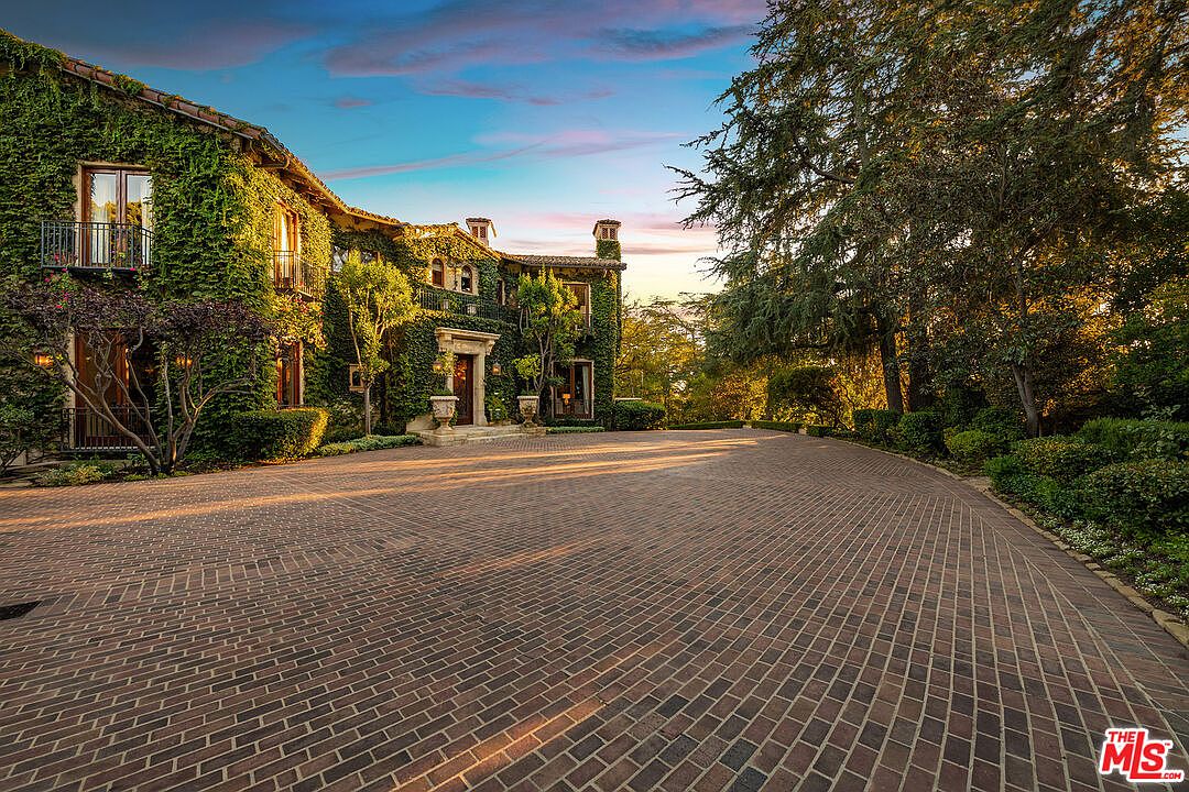 This grand, ivy-covered estate features a classic Mediterranean architectural style with a sweeping, curved brick driveway leading to the main entrance. The facade is adorned with multiple balconies and large windows, set against a backdrop of mature trees and a vibrant sunset sky. The perspective is a wide-angle shot from the driveway, emphasizing the impressive scale and curb appeal of the property.