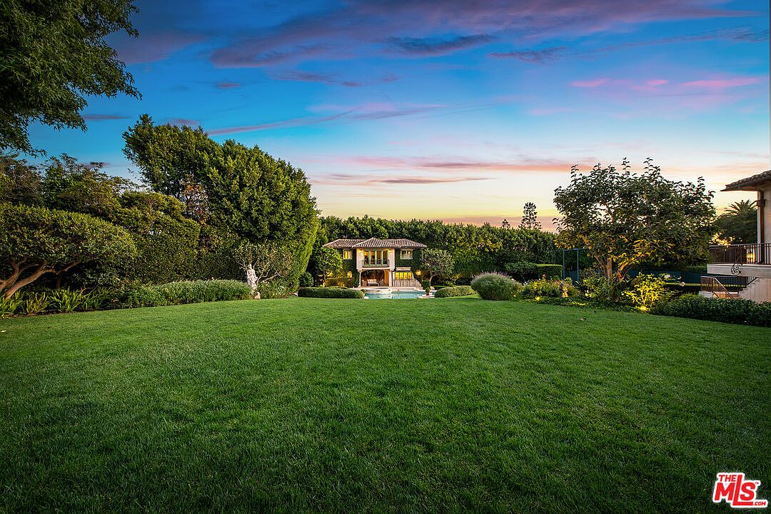 This expansive, manicured lawn serves as the foreground for a serene backyard scene, framed by lush, mature hedges and trees. In the distance, a two-story residence with a classic architectural style overlooks a swimming pool area, creating a private and tranquil atmosphere. The scene is captured during the golden hour, with a vibrant, colorful sunset sky casting a warm glow over the entire property.