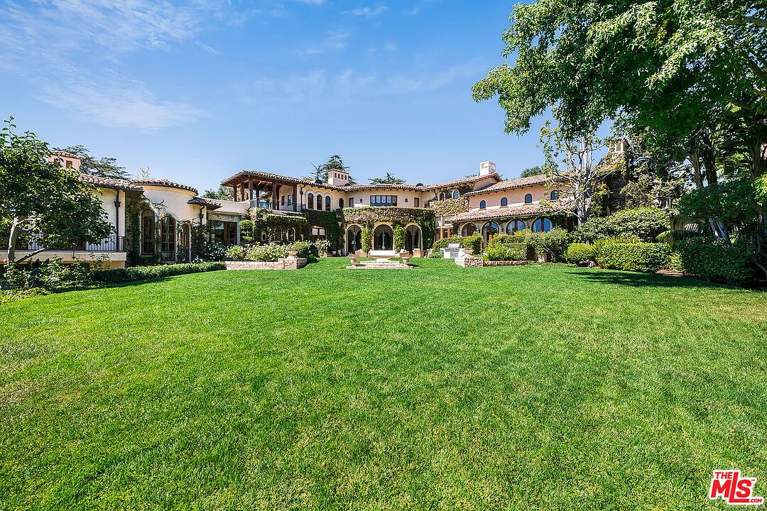 This expansive rear view of a Mediterranean-style estate showcases a sprawling, manicured lawn leading up to a multi-level residence with warm-toned stucco walls and a terracotta tile roof. The architecture features numerous arched windows, covered loggias, and climbing greenery that softens the facade, creating a grand and inviting atmosphere. A large, mature tree frames the right side of the composition, providing a sense of scale and natural beauty to the property.