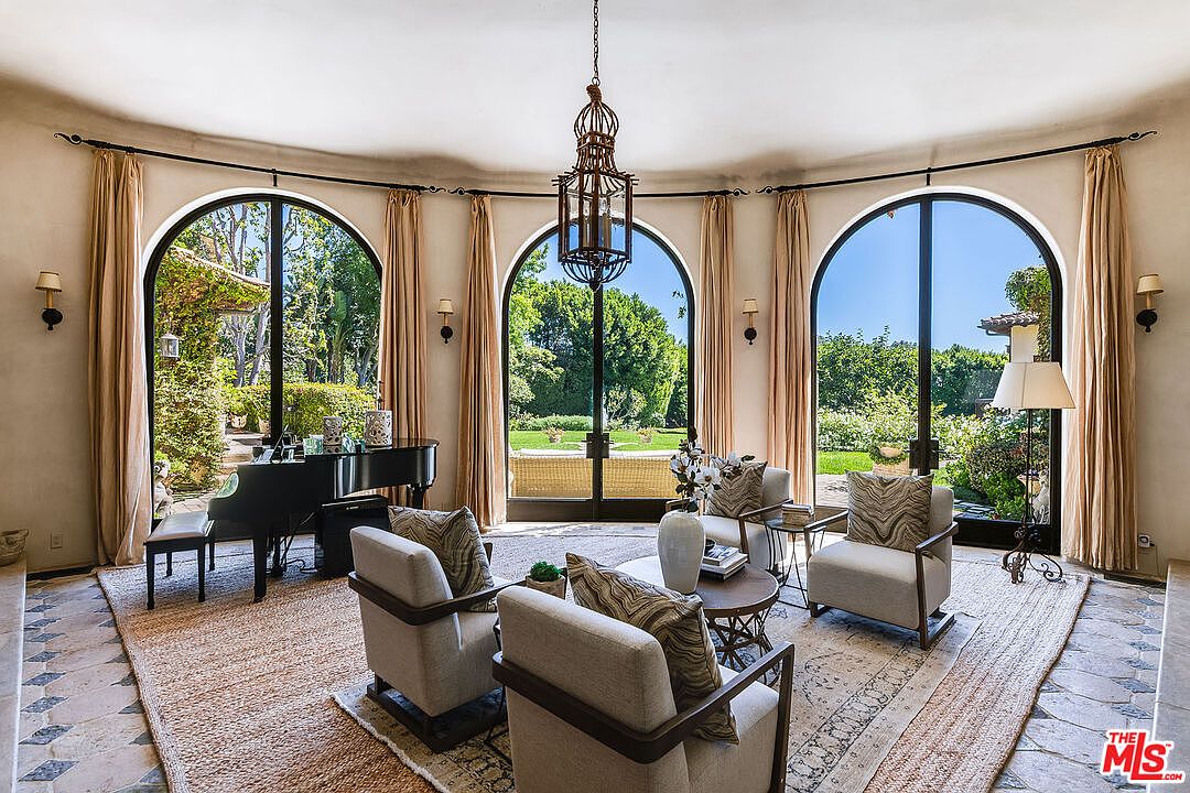 This elegant living room features a curved wall with three large arched windows that provide abundant natural light and views of the lush garden. The space is furnished with four upholstered armchairs arranged around a circular coffee table, with a grand piano positioned to the side. The room is anchored by a textured area rug over patterned tile flooring, all illuminated by a decorative hanging lantern fixture.