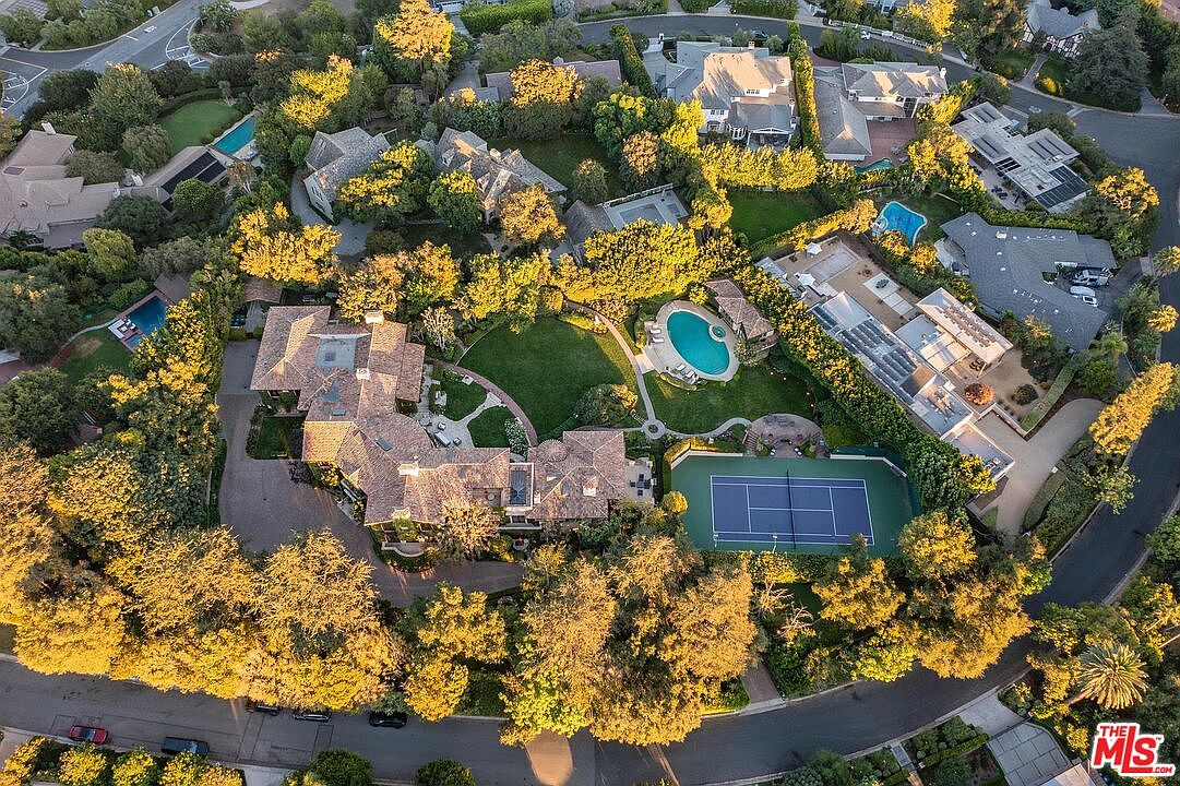 This high-angle aerial view captures a sprawling luxury estate nestled within a lush, tree-lined neighborhood. The property features a large, multi-winged residence with a tiled roof, a private tennis court, and a swimming pool set amidst manicured lawns and mature landscaping. The perspective highlights the expansive layout of the grounds and the seamless integration of outdoor recreational spaces.