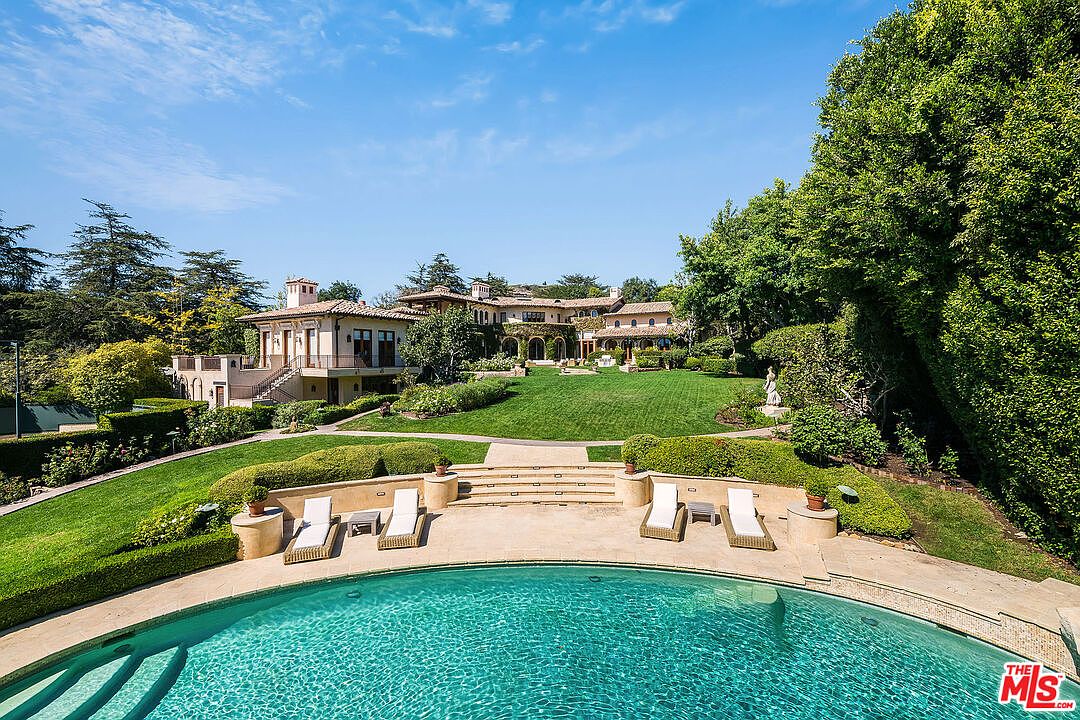 This elevated perspective captures a sprawling Mediterranean-style estate featuring a large, curved swimming pool in the foreground. Beyond the pool, a stone-paved terrace with lounge chairs leads to a lush, manicured lawn that slopes gently toward the multi-level residence. The home showcases classic architectural details, including a tiled roof and arched openings, set against a backdrop of mature trees and a clear blue sky.