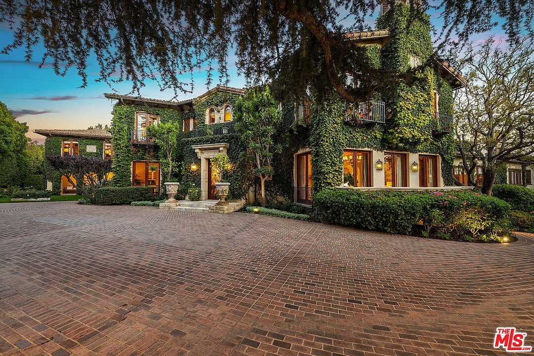 This grand, ivy-covered Mediterranean-style estate features a sprawling brick-paved motor court leading to a prominent arched entryway. The multi-story facade is adorned with lush climbing greenery, wrought-iron balcony railings, and warm exterior lighting that highlights the architectural details. The perspective is a wide-angle shot from the driveway, capturing the imposing scale and mature landscaping of the property at dusk.