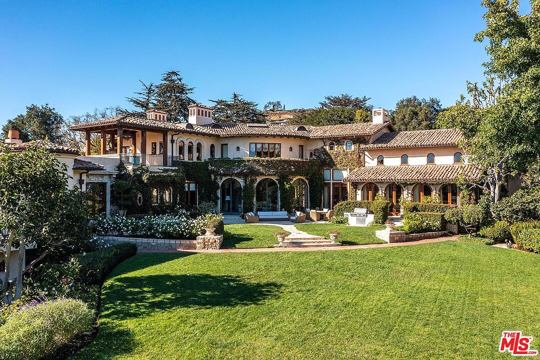 This expansive Mediterranean-style estate features a multi-level rear facade with warm-toned stucco walls, terracotta roof tiles, and climbing ivy. The home opens onto a manicured lawn and a stone-paved patio area, complete with outdoor seating and a built-in grill station. The perspective captures the grand scale of the residence, emphasizing its seamless integration with the lush, landscaped grounds and mature trees.
