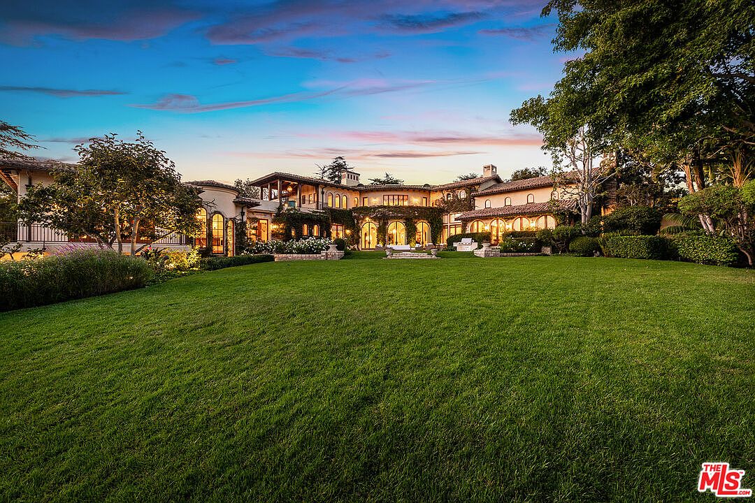 This expansive Mediterranean-style estate is captured from a low-angle perspective across a vast, manicured lawn. The rear facade features warm-toned stucco walls, arched windows, and a classic clay tile roof, with lush greenery and climbing vines softening the architectural lines. The scene is bathed in the golden light of dusk, creating an inviting and luxurious atmosphere.