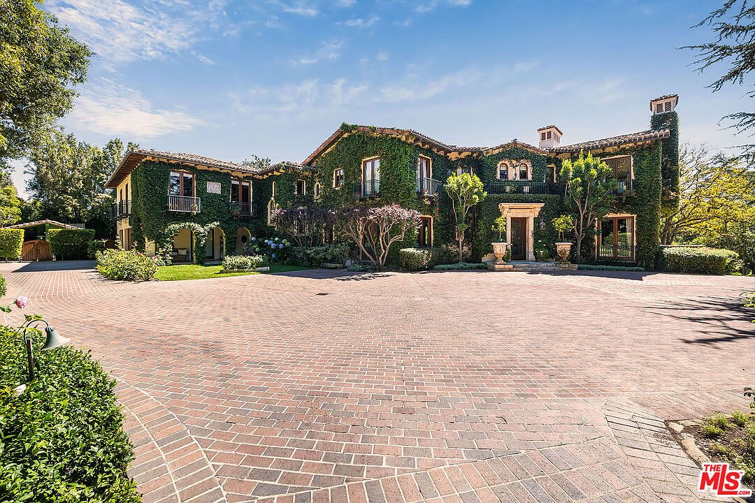 This grand, ivy-covered Mediterranean-style estate features a sprawling brick-paved motor court leading to a prominent arched entryway. The multi-story residence is characterized by its lush greenery, classic architectural details, and symmetrical facade, creating an impression of timeless elegance and privacy. The perspective is a wide-angle shot from the driveway, capturing the full scale of the home against a clear blue sky.