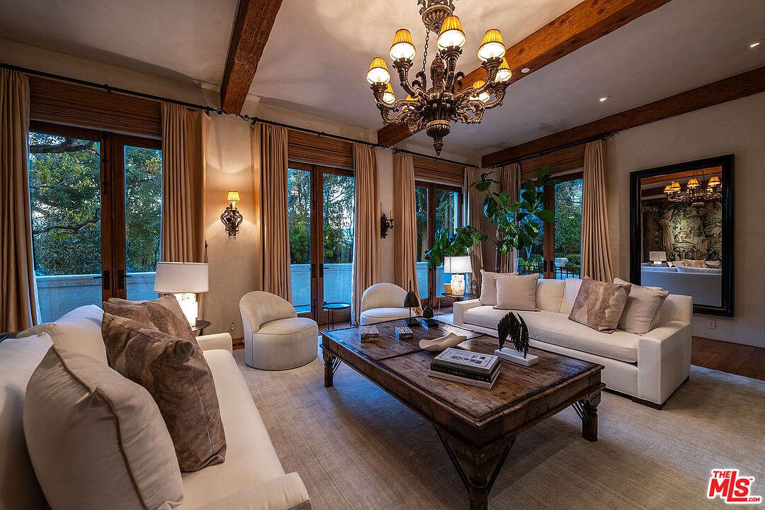 This elegant living room features a sophisticated neutral color palette, highlighted by a large, rustic wooden coffee table centered between two plush white sofas and two matching armchairs. The space is illuminated by a grand, multi-tiered chandelier suspended from a ceiling accented with exposed wooden beams, while large glass doors provide ample natural light and views of the surrounding greenery. A large, framed mirror on the wall reflects the room's refined decor, creating an inviting and luxurious atmosphere.