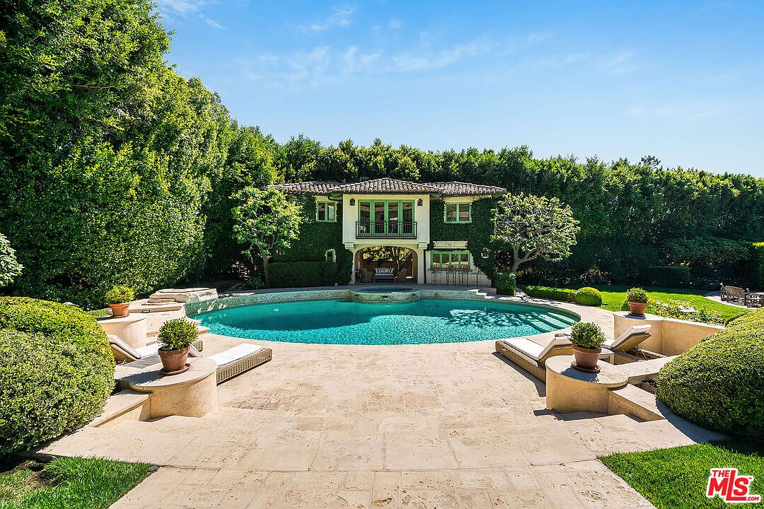 This image captures a serene backyard oasis featuring a large, curved swimming pool as the central focal point. The pool area is surrounded by a light-colored stone patio, lounge chairs, and lush, manicured greenery that provides significant privacy. In the background, a two-story structure covered in climbing ivy features a balcony and a covered patio area, creating a sophisticated and tranquil retreat.