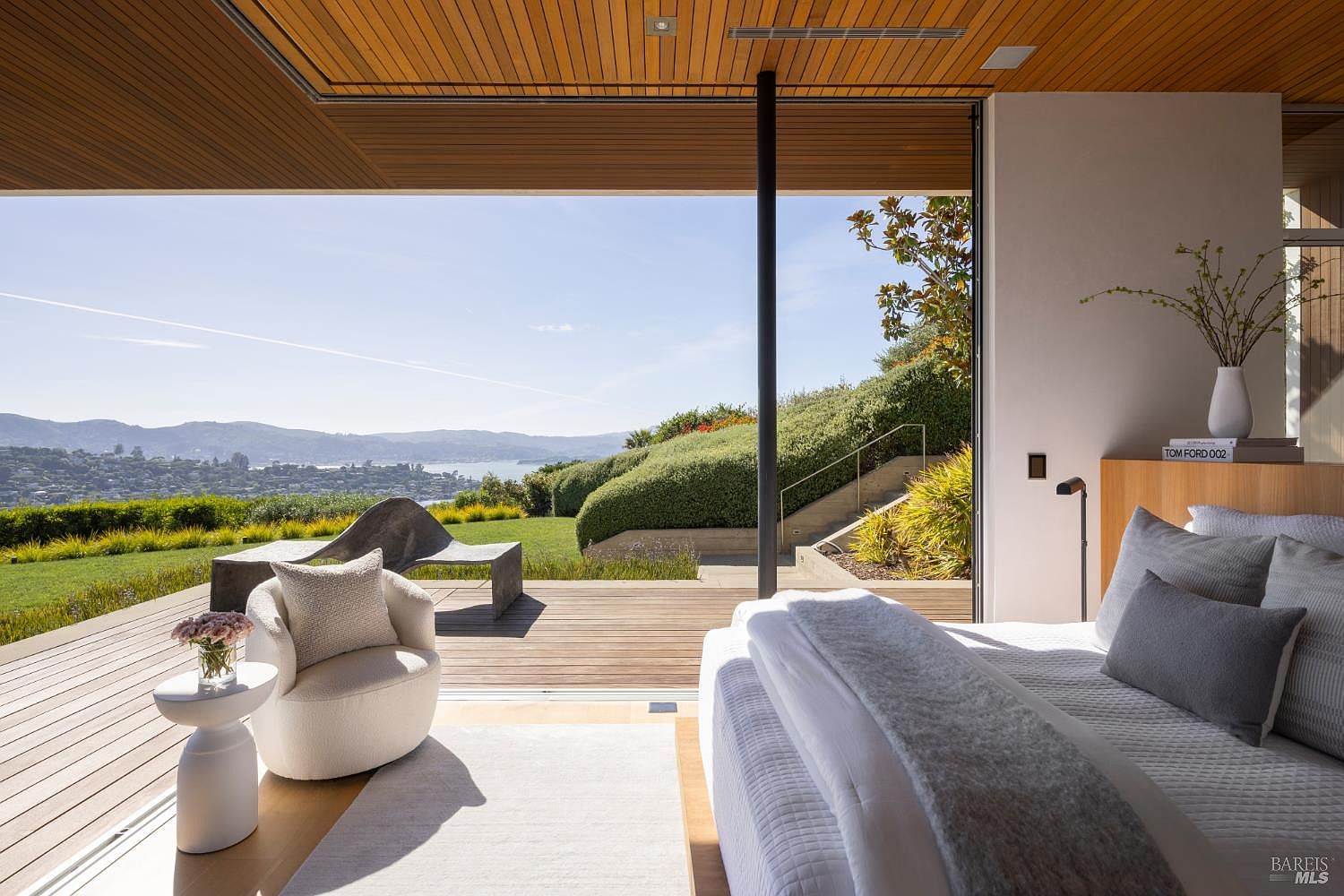 This primary bedroom features a seamless transition to an expansive outdoor deck through large sliding glass doors, framing a panoramic view of the bay and rolling hills. The interior is characterized by warm wood-paneled ceilings, a plush bed with neutral linens, and a cozy armchair placed near the window to capture the scenery. The overall aesthetic is serene and sophisticated, emphasizing a strong connection between indoor living and the natural landscape.