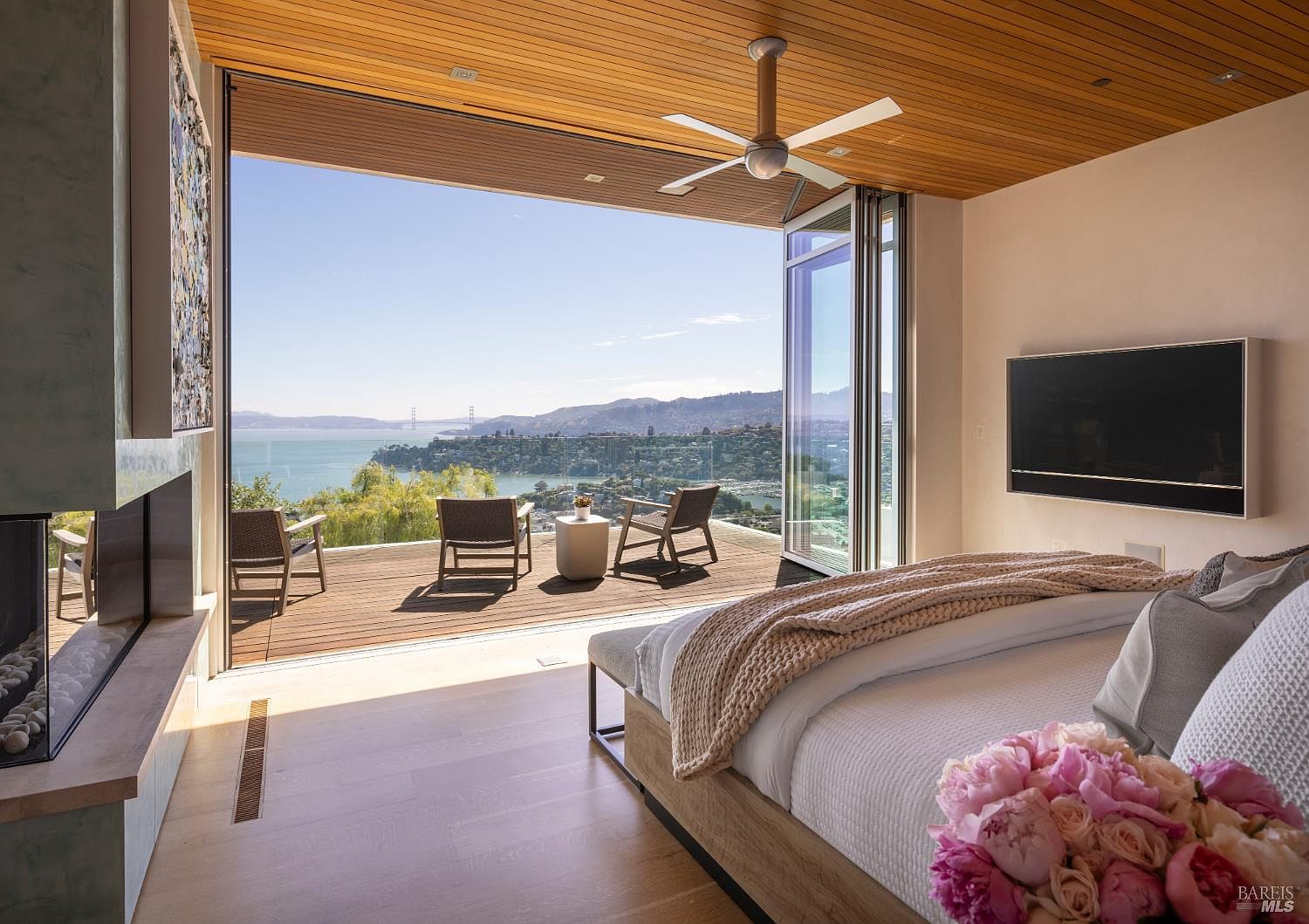This luxurious primary bedroom features a seamless transition to an expansive outdoor deck through large folding glass doors, framing a panoramic view of the bay and distant bridge. The room is appointed with light wood flooring, a modern ceiling fan, and a wall-mounted television, while a bouquet of pink peonies adds a soft touch to the foreground. The aesthetic is contemporary and serene, emphasizing the connection between the indoor living space and the natural landscape.