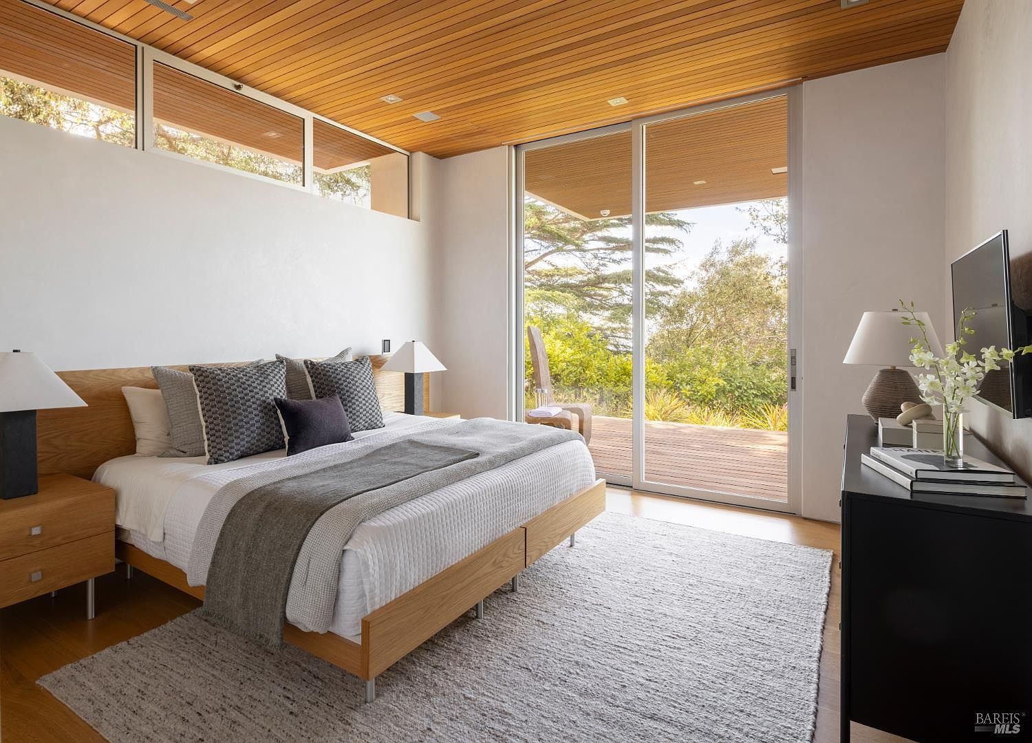 This primary bedroom features a warm, modern aesthetic with a light wood-paneled ceiling and a matching headboard. A large sliding glass door provides direct access to an outdoor deck, filling the room with natural light and offering a serene view of the surrounding trees. The space is furnished with a neutral-toned bed, matching nightstands, and a sleek black dresser, creating a calm and sophisticated atmosphere.