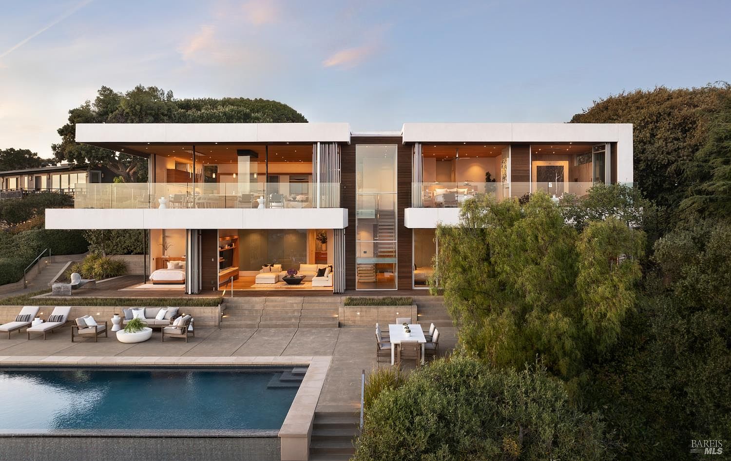 This modern, two-story luxury residence features a clean, geometric design with extensive glass walls that blur the line between indoor and outdoor living. The rear exterior showcases a spacious concrete patio, a rectangular swimming pool, and an outdoor dining area, all set against a backdrop of mature trees. The home's architecture emphasizes horizontal lines, warm wood accents, and a central glass-enclosed staircase that serves as a striking focal point.