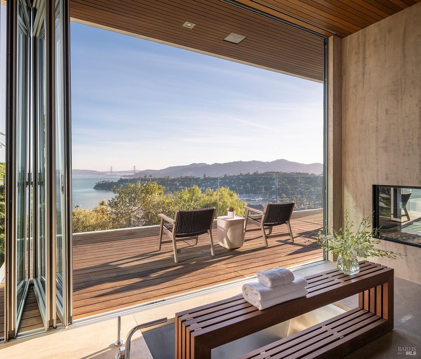 This luxurious bathroom space features a seamless transition to an outdoor deck through expansive folding glass doors, offering a panoramic view of the bay and distant bridge. The interior includes a deep soaking tub positioned next to a wooden bench with folded towels, while the deck is furnished with two chairs and a small table. The design emphasizes a sophisticated indoor-outdoor lifestyle with clean lines and a serene, high-end aesthetic.