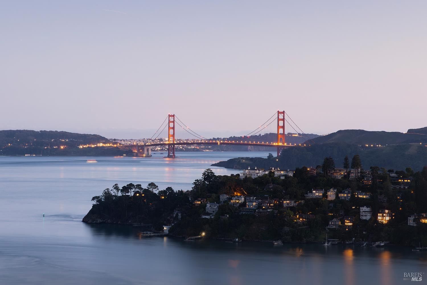 This wide-angle aerial view captures a serene twilight scene overlooking a coastal residential area with the iconic Golden Gate Bridge spanning the horizon. The foreground features a lush, tree-lined hillside dotted with illuminated homes, leading down to the calm waters of the bay. The composition emphasizes the prestigious location and the dramatic, expansive perspective of the San Francisco Bay landscape.