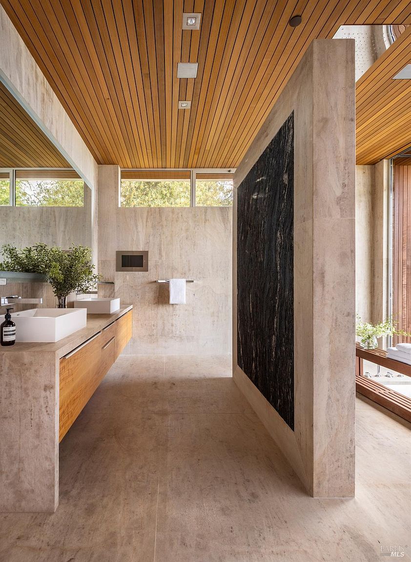 This luxurious primary bathroom features a minimalist, spa-inspired design with warm wood-slat ceilings and expansive stone surfaces. A floating vanity with dual sinks is positioned opposite a striking, dark stone-clad partition wall that separates the vanity area from the shower space. The room is bathed in natural light from high-set windows, creating an airy and sophisticated atmosphere.