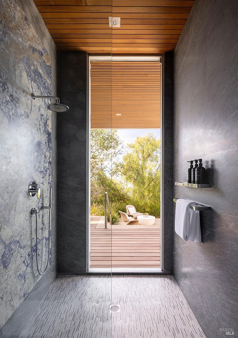 This modern walk-in shower features a striking blue-veined stone accent wall contrasted by dark, textured side walls and a light-toned linear tile floor. A floor-to-ceiling glass panel provides an unobstructed view through the shower to an outdoor deck area, creating a seamless indoor-outdoor connection. The space is finished with a warm wood-slat ceiling, a wall-mounted shower head, and a sleek shelf for toiletries.