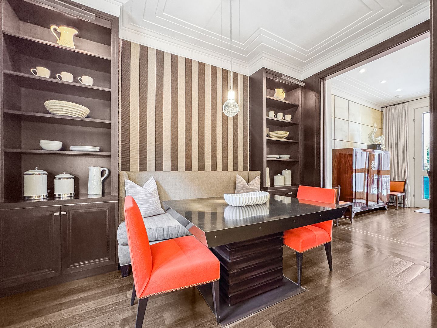 This elegant dining nook features a built-in banquette set against a wall with bold, vertical-striped wallpaper, flanked by dark wood shelving units. A modern, dark-toned table is paired with two vibrant orange chairs, creating a striking contrast in the space. The room is illuminated by a single pendant light, and the perspective looks through an open doorway into an adjacent room with a polished wood cabinet.
