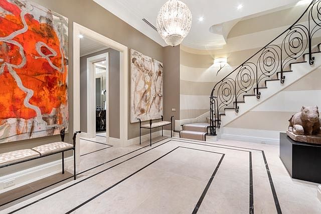 This elegant grand foyer features a sweeping staircase with intricate wrought-iron railings and light-toned stone flooring accented by dark inlaid borders. The space is adorned with large, vibrant abstract artwork and a sophisticated crystal chandelier, creating a luxurious and welcoming atmosphere. A view through the doorway reveals a glimpse into an adjacent room, emphasizing the open and airy layout of the home.