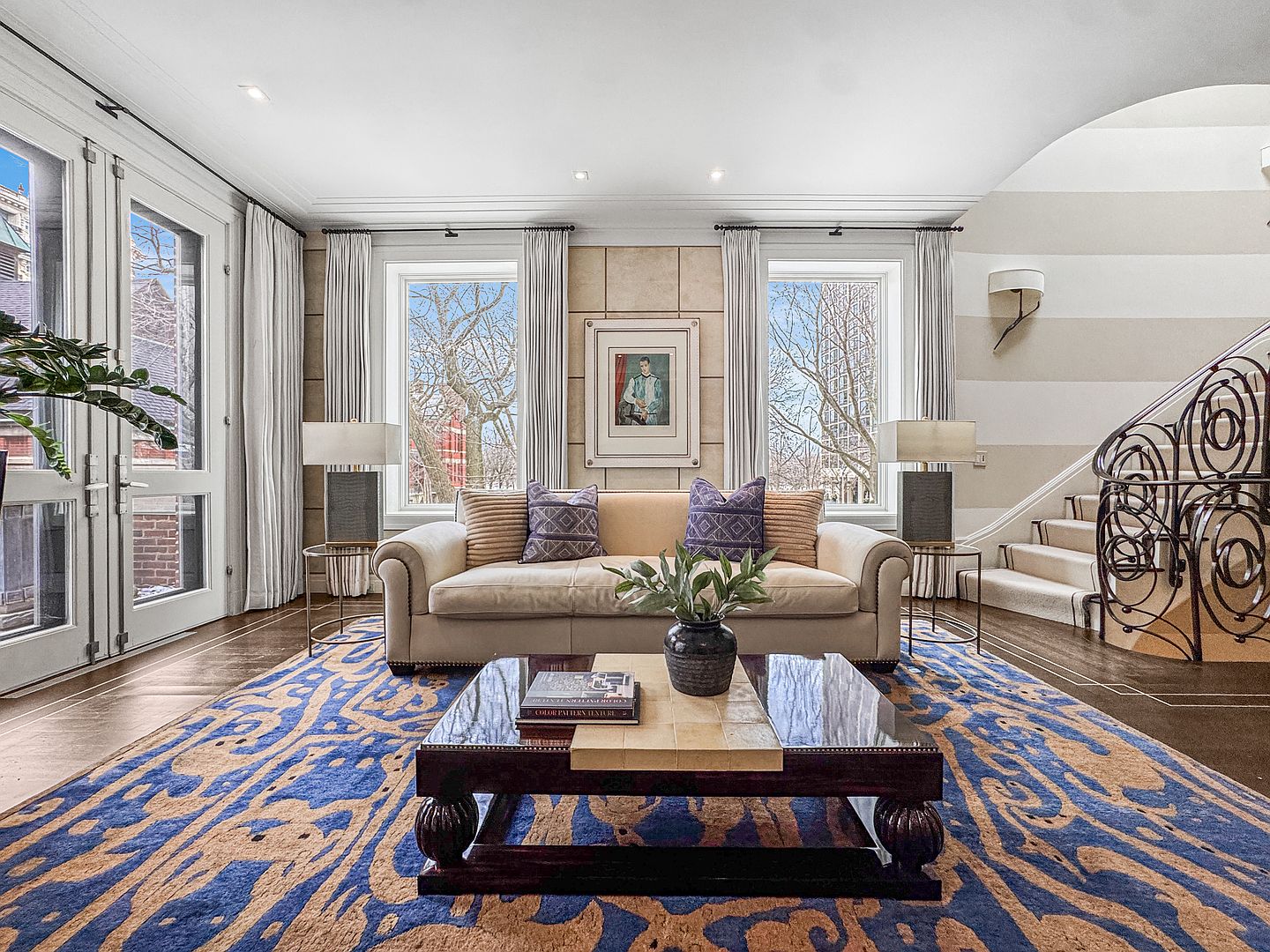 This elegant living room features a neutral-toned sofa centered between two large windows that offer views of the outdoors. A bold, blue and tan patterned area rug anchors the space, complemented by a dark wood coffee table and a decorative framed portrait on the wall. To the right, a graceful staircase with an ornate metal railing leads upward, while the room is finished with soft drapery and warm wall sconces.