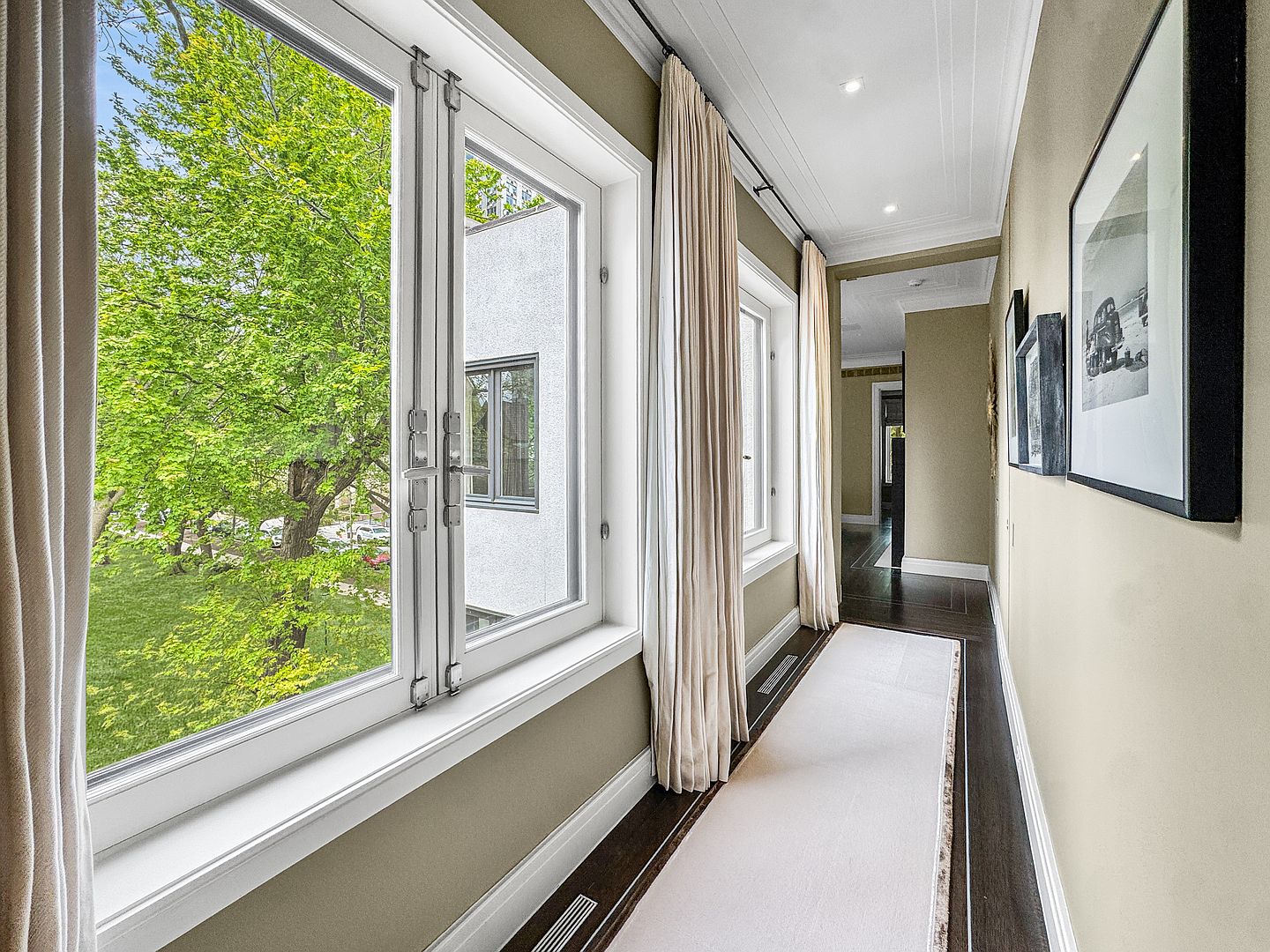 This elegant hallway features dark wood flooring topped with a long, light-colored runner, creating a sophisticated contrast. Large windows with cream-colored curtains allow ample natural light to fill the space, offering a view of lush green trees outside. The walls are painted in a neutral tone and adorned with framed black-and-white artwork, leading the eye toward the transition into the next room.