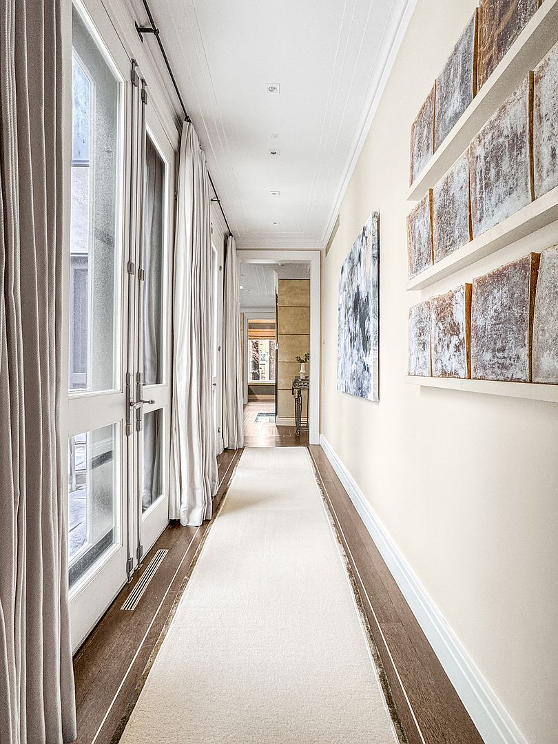 This elegant, light-filled hallway features a long runner rug over dark wood flooring, leading the eye toward a distant doorway. To the left, tall glass doors with white frames are draped in soft, neutral curtains, while the right wall is adorned with a series of framed, textured art pieces. The space is finished with a clean, neutral color palette and recessed ceiling lighting, creating a sophisticated and airy atmosphere.