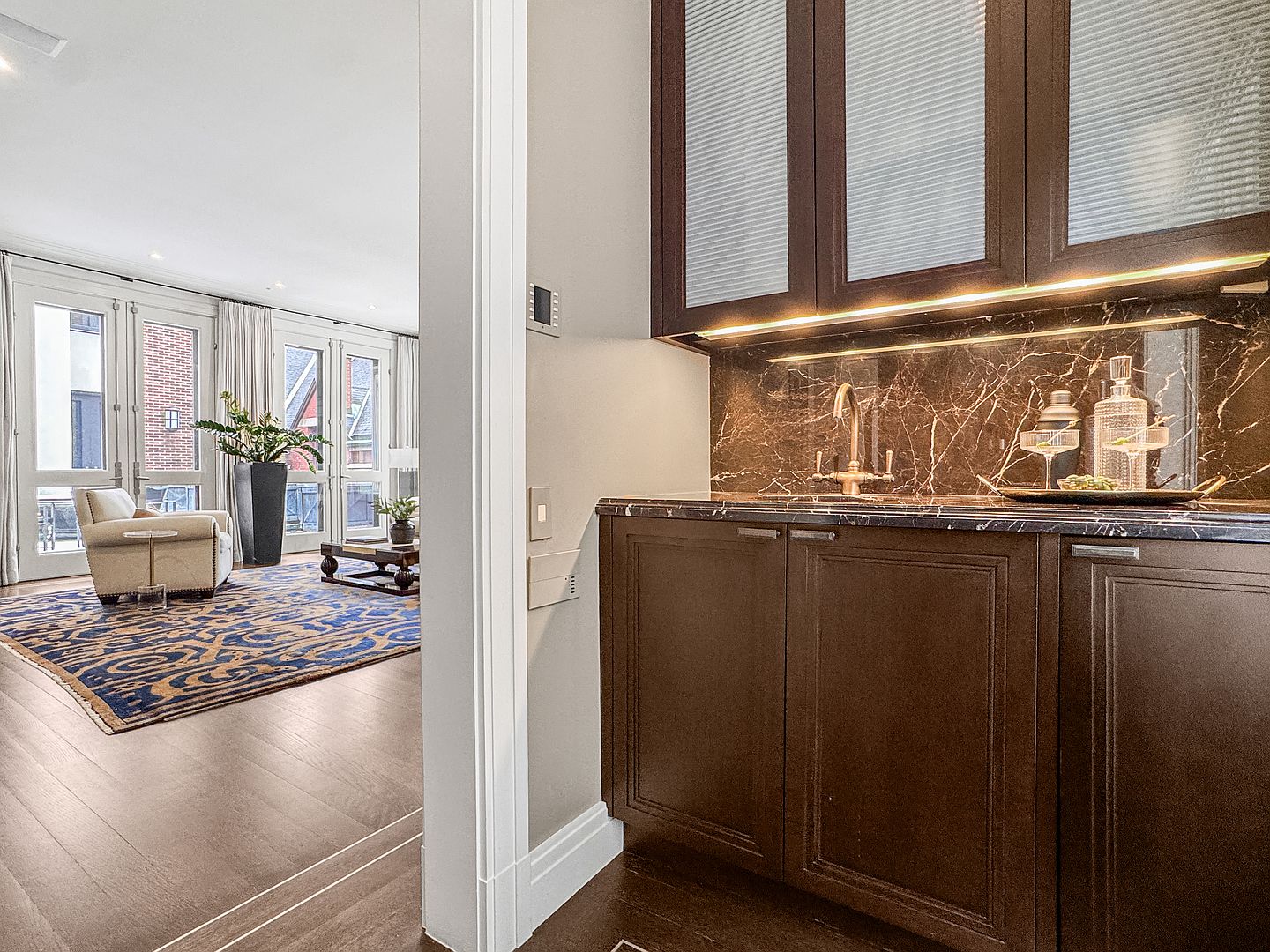 This image captures a sophisticated wet bar area featuring dark cabinetry with glass-paneled upper doors and a stone backsplash. The foreground highlights a polished stone countertop with a small sink, while the background reveals a bright, open living space with large windows and a patterned area rug. The perspective transitions from the intimate, moody bar area into the airy, sunlit living room, creating a sense of depth and luxury.