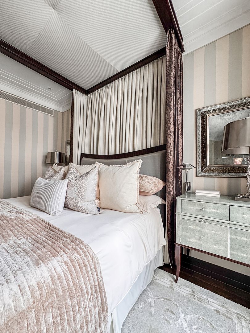 This elegant primary bedroom features a sophisticated four-poster bed draped with soft, neutral-toned curtains and layered with plush, textured bedding. The room is accented by striped wallpaper, a decorative mirror above a textured bedside chest, and a patterned area rug, creating a refined and tranquil atmosphere. The perspective is a close-up, eye-level shot that highlights the luxurious textures and classic design elements of the space.