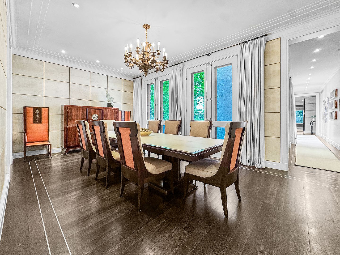 This elegant dining room features a large rectangular table surrounded by eight high-backed chairs with orange upholstered accents. The space is anchored by a dark wood sideboard against a wall with a grid-patterned panel design, while a classic chandelier hangs above the table. Tall glass doors with white frames lead to an outdoor area, allowing natural light to fill the room, which is finished with dark wood flooring and a long hallway visible in the background.
