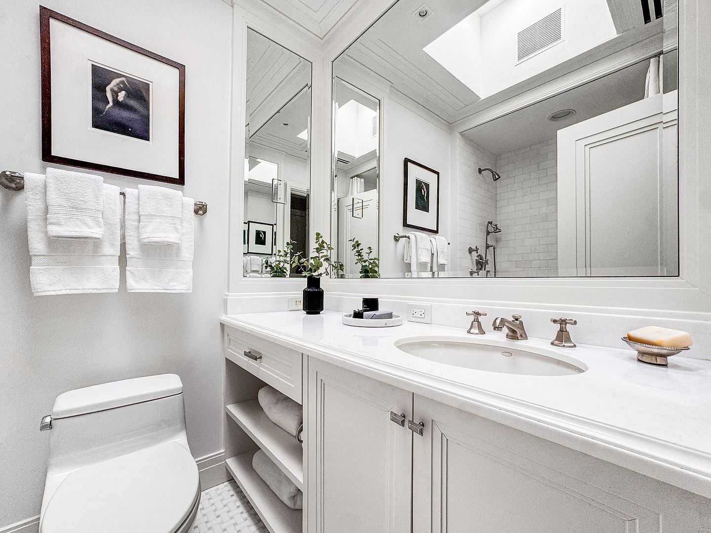 This bright and clean bathroom features a white vanity with a stone countertop, an integrated sink, and metallic fixtures. A large mirror spans the wall above the vanity, reflecting the shower area and a framed piece of art, while open shelving provides convenient storage for towels. The space is finished with a white toilet and patterned floor tile, creating a cohesive and polished aesthetic.