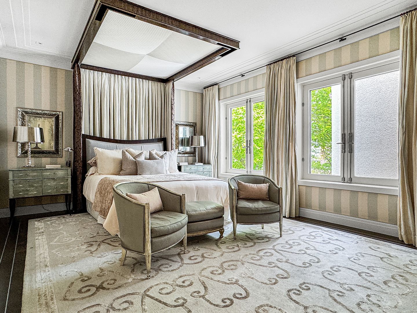 This elegant primary bedroom features a grand canopy bed as the focal point, framed by soft drapery and a dark wood structure. The room is decorated with a sophisticated neutral palette, including striped wallpaper, a large patterned area rug, and two upholstered armchairs with an ottoman. Large windows with floor-length curtains allow natural light to fill the space, creating a serene and luxurious atmosphere.