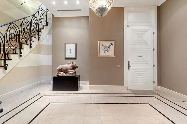 This elegant foyer features a grand, curved staircase with intricate black wrought-iron railings and light-toned stone flooring with a decorative dark border. A central pedestal displays a stone animal sculpture, flanked by two framed artworks on the neutral-toned walls. The space is illuminated by a decorative chandelier, creating a sophisticated and welcoming entrance.