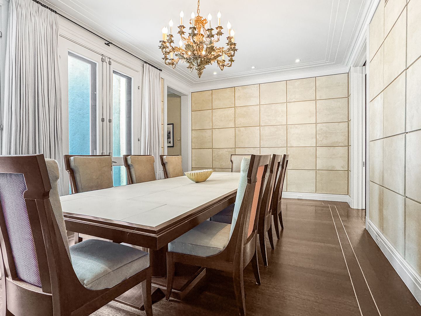 This elegant dining room features a long, light-toned table surrounded by upholstered chairs with dark wood frames. A grand, ornate gold chandelier serves as the focal point above the table, while the walls are adorned with a grid-patterned, textured paneling. The room is brightened by tall windows draped in floor-to-ceiling curtains, creating a sophisticated and formal atmosphere.