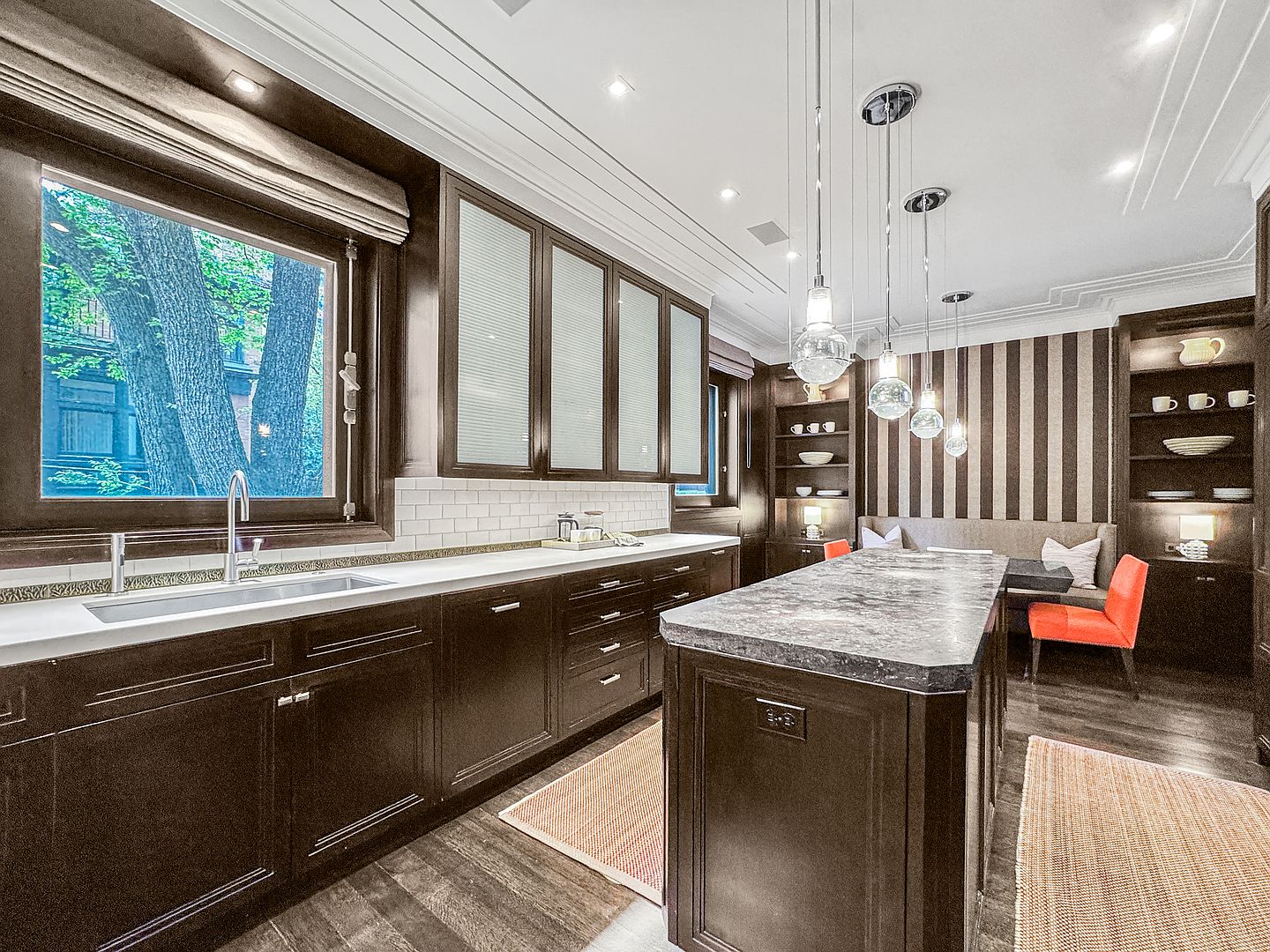 This sophisticated kitchen features dark wood cabinetry paired with light stone countertops and a matching central island. A breakfast nook with striped wallpaper and vibrant orange chairs adds a pop of color, while three glass pendant lights hang above the island. The space is framed by a large window overlooking trees, creating a warm and inviting atmosphere.