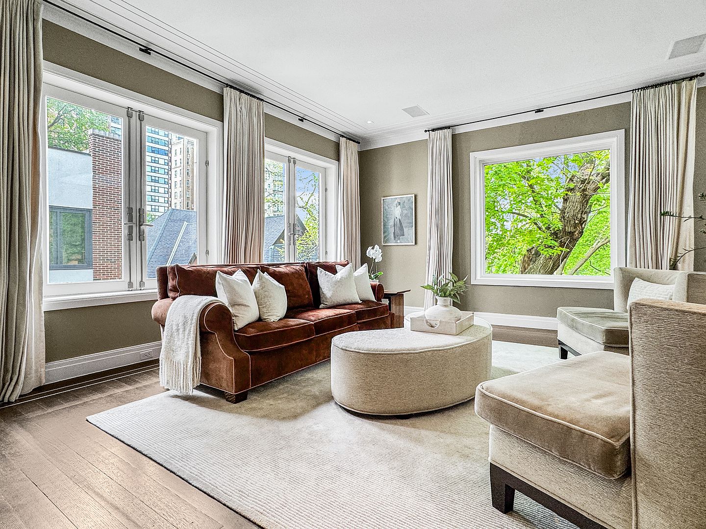 This elegant living room features a rich brown velvet sofa accented with white throw pillows, centered around a large, light-toned oval ottoman. The space is anchored by a neutral area rug over light wood flooring, with tall windows draped in floor-length curtains that allow for abundant natural light. A pair of upholstered armchairs completes the seating arrangement, while a framed piece of art on the wall adds a sophisticated touch to the serene, neutral-toned interior.
