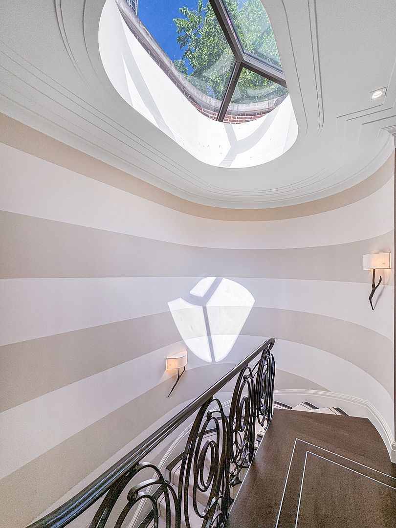 This elegant staircase features a curved wall adorned with horizontal stripes in neutral tones, leading up to a striking oval skylight that floods the space with natural light. A decorative, dark metal railing with intricate scrollwork follows the curve of the stairs, which are covered in a dark, plush carpet. Two wall-mounted sconces provide soft, warm accent lighting, enhancing the sophisticated and architectural character of the entryway.